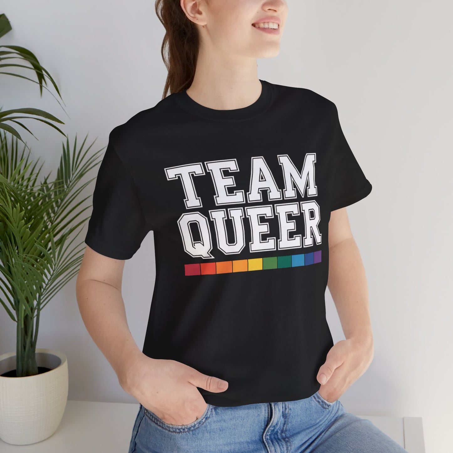 Person wearing a black t-shirt with 'TEAM QUEER' text and rainbow stripes, standing indoors.