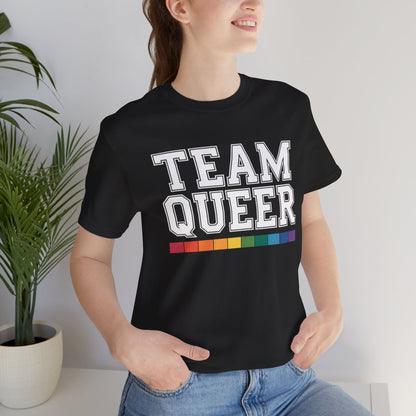 Person wearing a black t-shirt with 'TEAM QUEER' text and rainbow stripes, standing indoors.