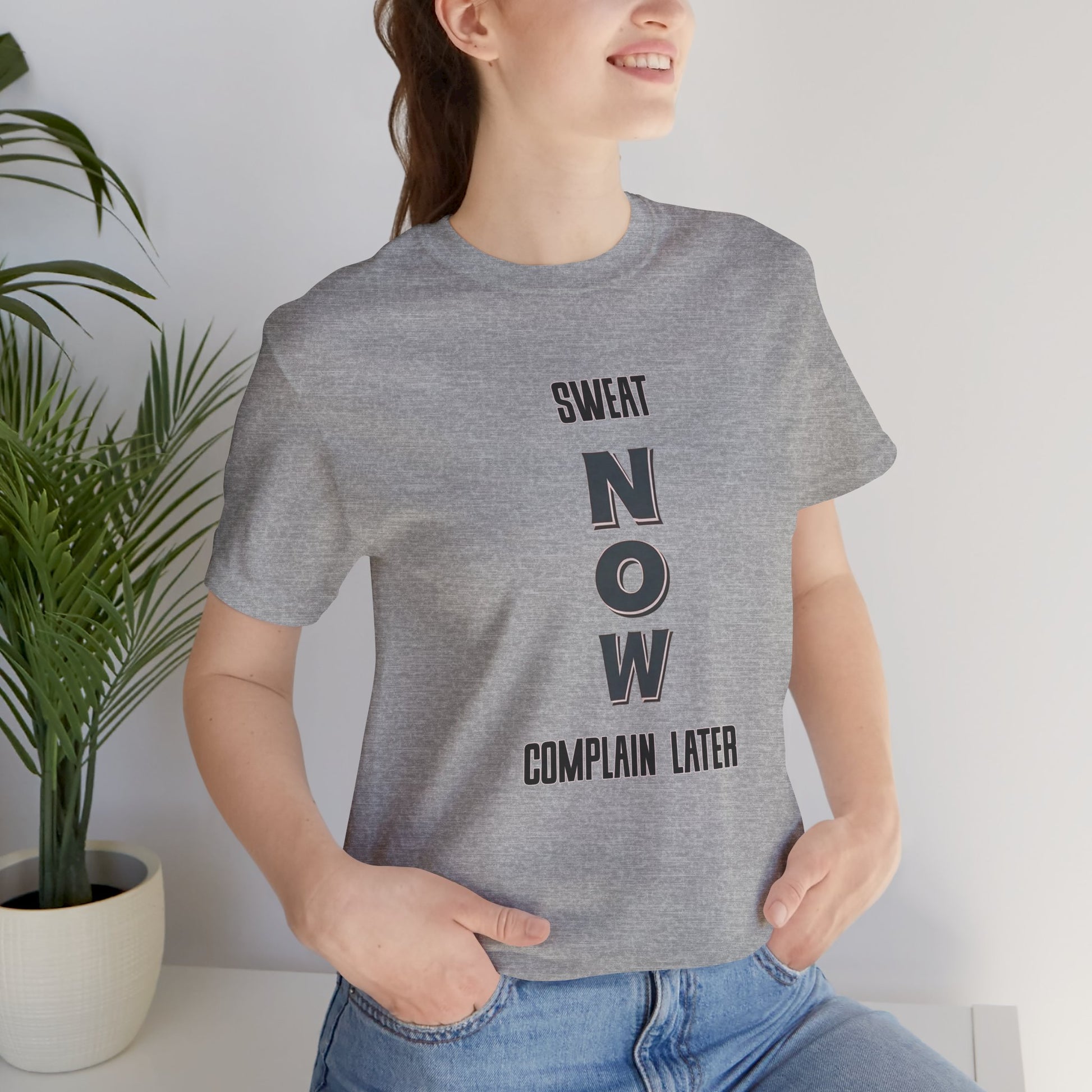 Person wearing a gray t-shirt with 'SWEAT NOW COMPLAIN LATER' text, standing indoors.