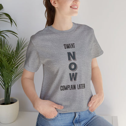 Person wearing a gray t-shirt with 'SWEAT NOW COMPLAIN LATER' text, standing indoors.