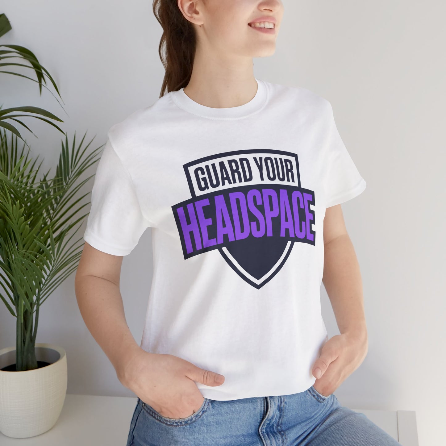 Person wearing a white t-shirt with 'Guard Your Headspace' text, standing indoors next to a plant.