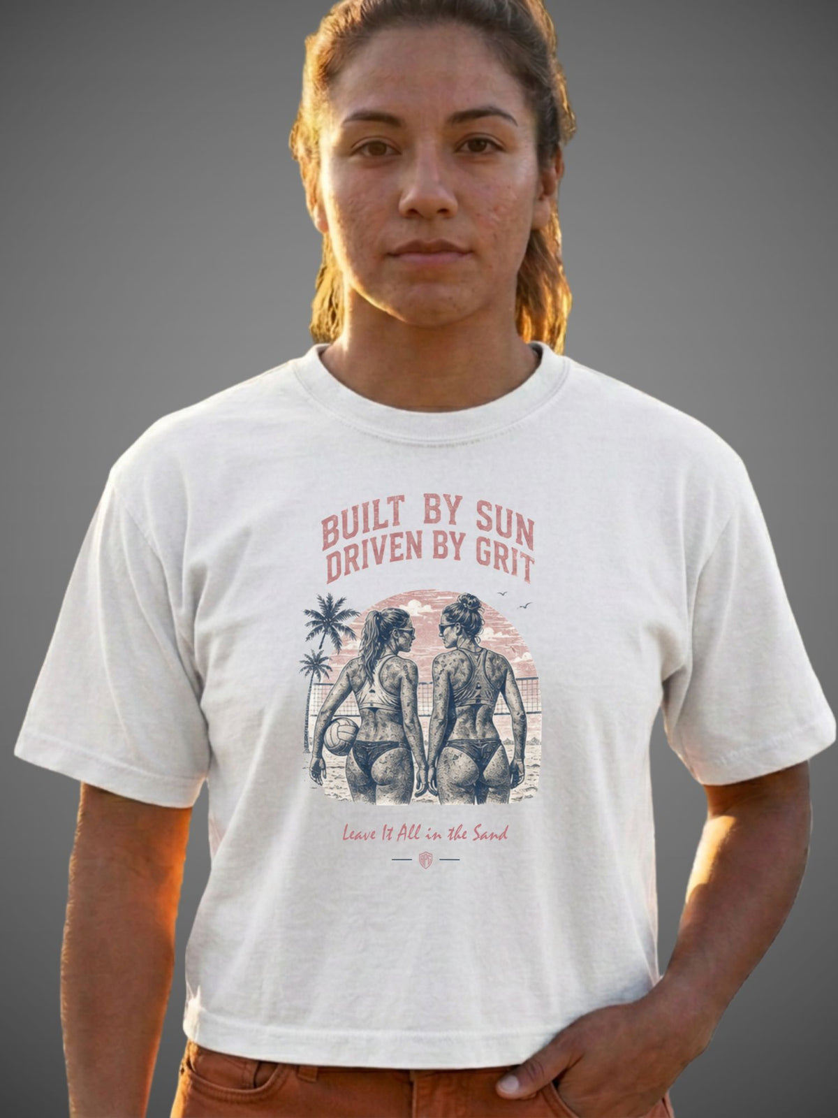A woman wearing a white graphic t-shirt with a vintage-style print of two women on a beach and the text 'BUILT BY SUN DRIVEN BY GRIT' and 'Leave It All in the Sand'.