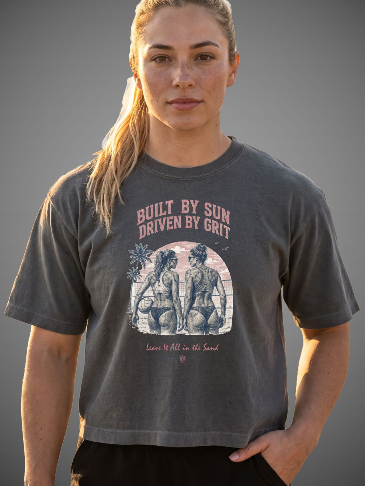 A woman modeling a dark gray graphic t-shirt with the text 'BUILT BY SUN DRIVEN BY GRIT' and a beach-themed illustration on the front, set against a solid gray background.