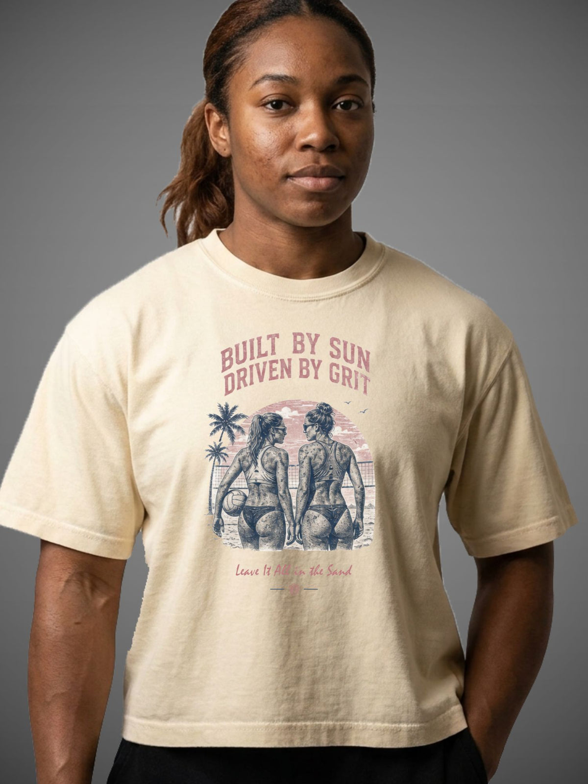 A woman wearing a cream-colored graphic t-shirt with a vintage-style print of two women on a beach and the text 'BUILT BY SUN DRIVEN BY GRIT'.