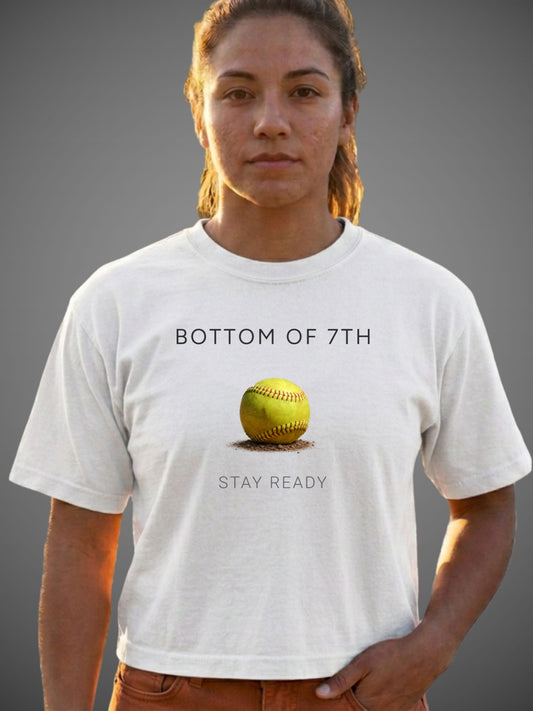 A woman wearing a white graphic t-shirt with a yellow softball graphic and the text 'BOTTOM OF 7TH' and 'STAY READY'.