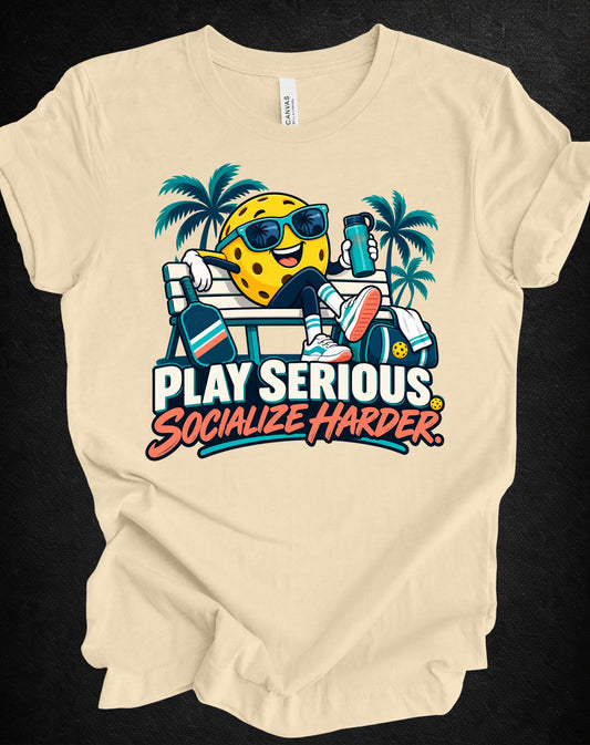 A beige graphic t-shirt laid flat on a dark textured background. The shirt features a colorful cartoon graphic of a yellow cheese character wearing sunglasses and sitting on a beach chair with palm trees, above the text 'PLAY SERIOUS.