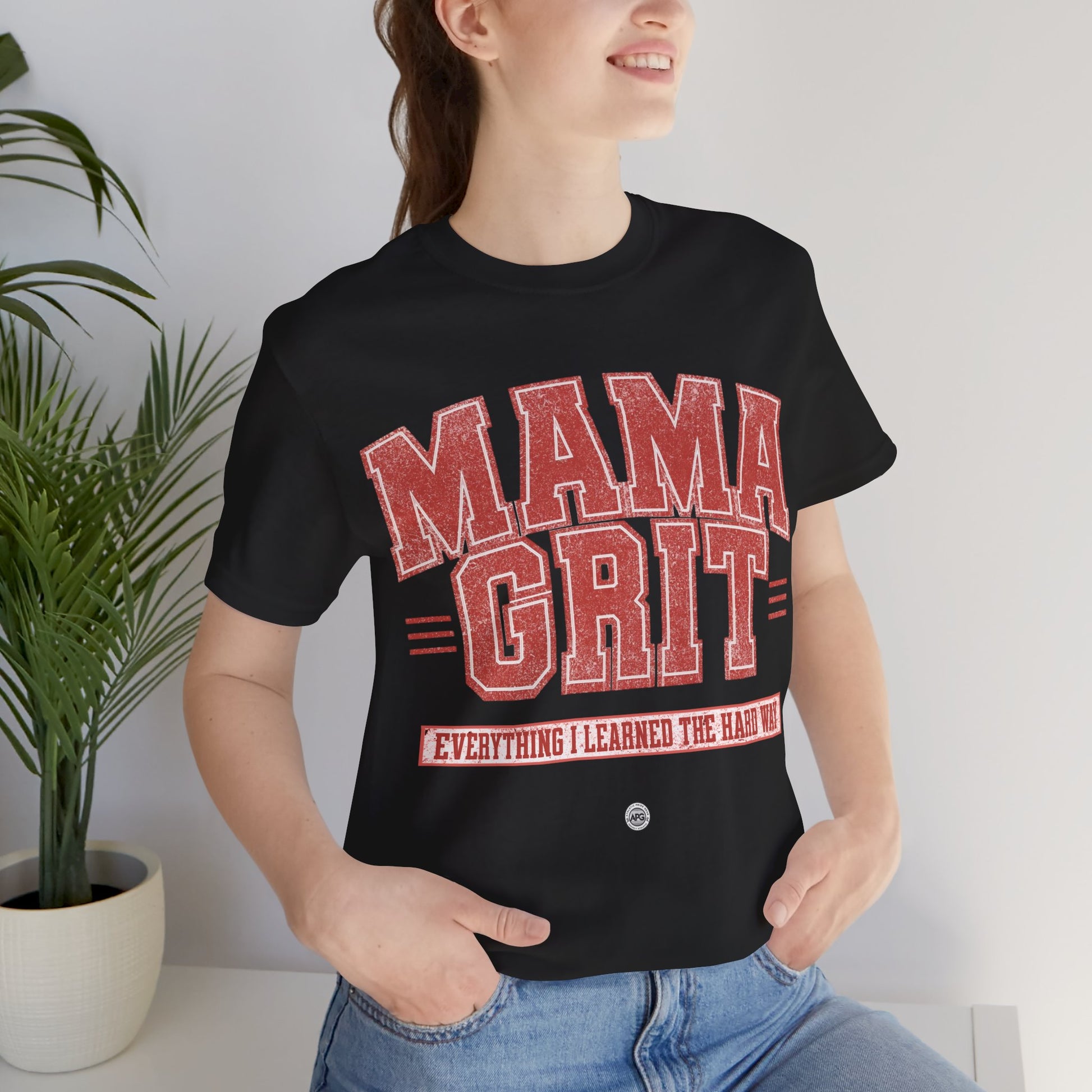 Person wearing a black t-shirt with 'MAMA GRIT' text, standing indoors next to a plant.