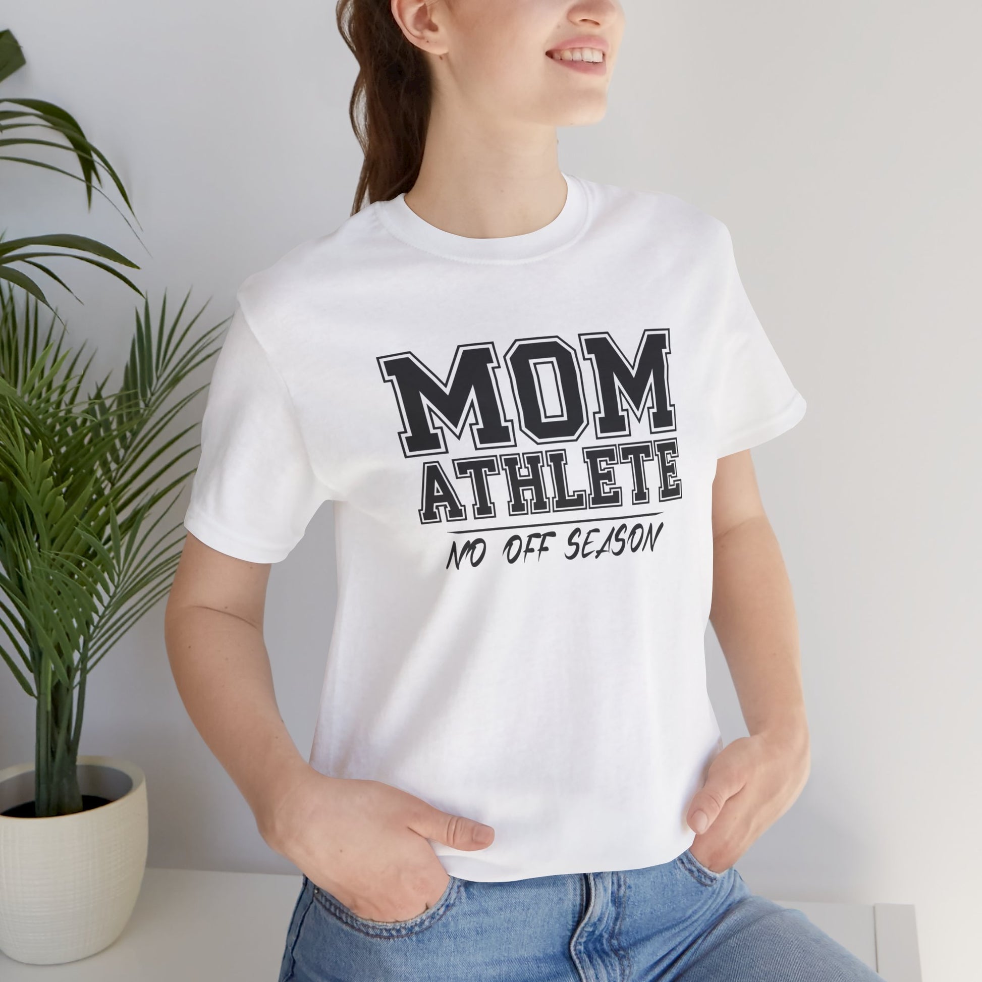 Person wearing a white t-shirt with 'MOM ATHLETE NO OFF SEASON' text, standing indoors next to a plant.
