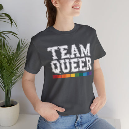 Person wearing a black t-shirt with 'TEAM QUEER' text and rainbow stripes, standing indoors.